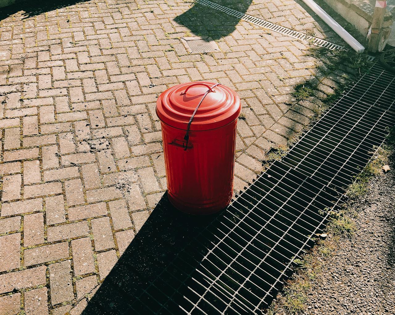 High angle of red metal trash can placed on pavement near sewer grates in city street in sunny summer day